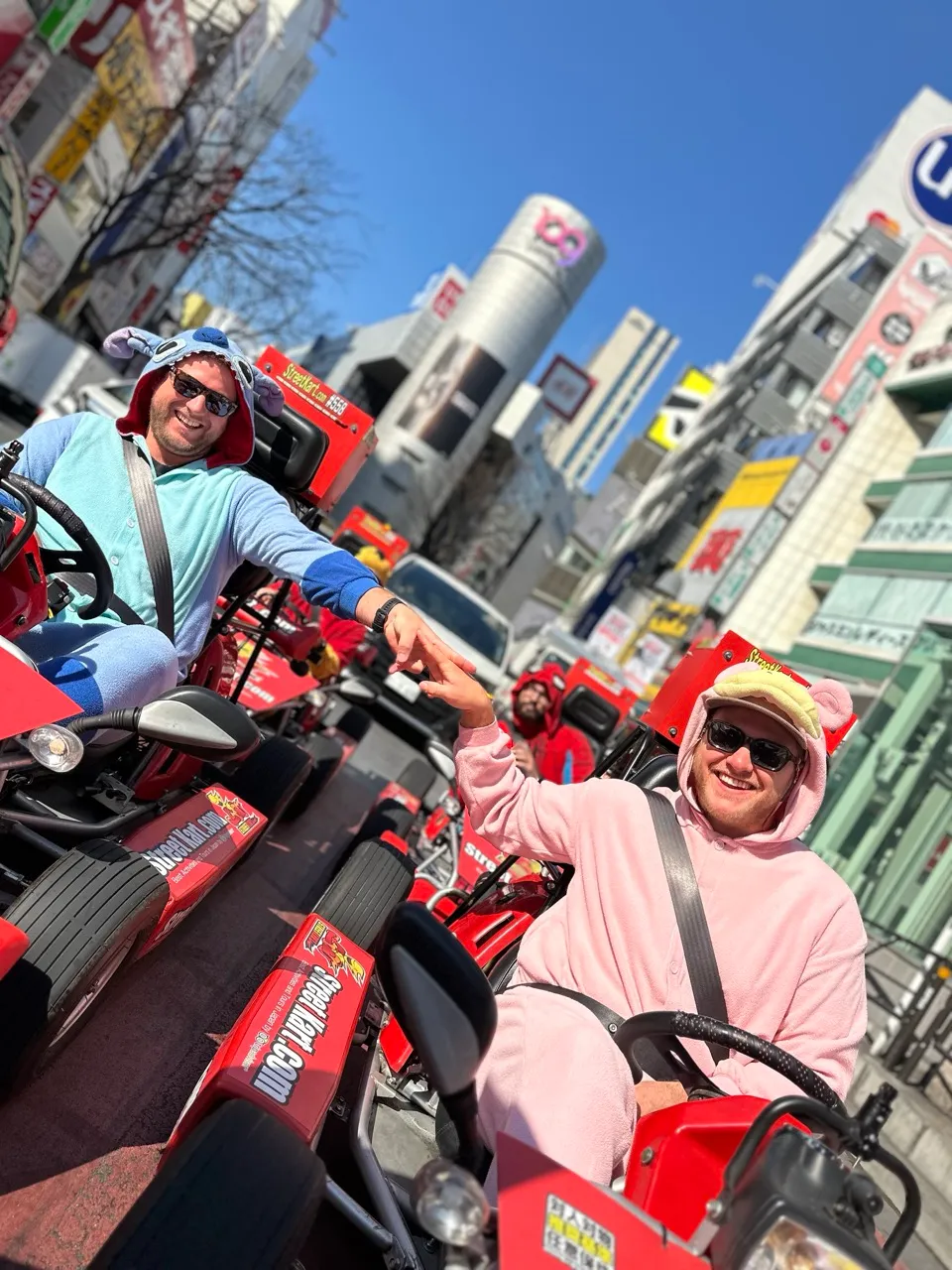 Two men in colorful hoodies ride red go-karts in a busy city street, high-fiving each other under a clear blue sky.
