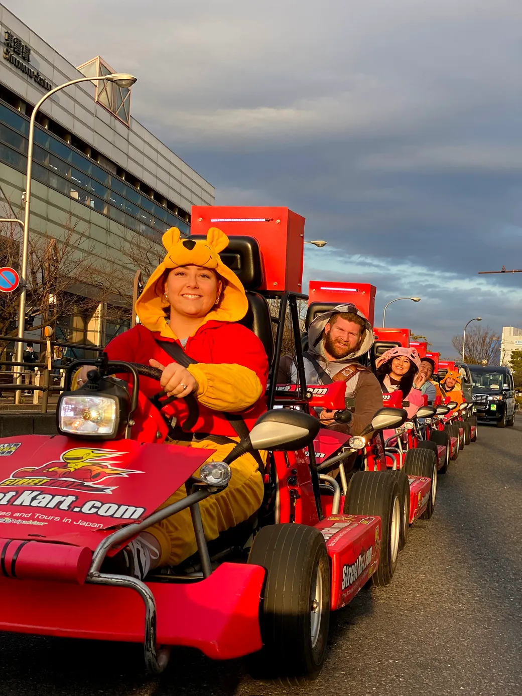 Woman in a yellow bear costume driving a red go-kart at a parade, with a line of similar karts behind her.
