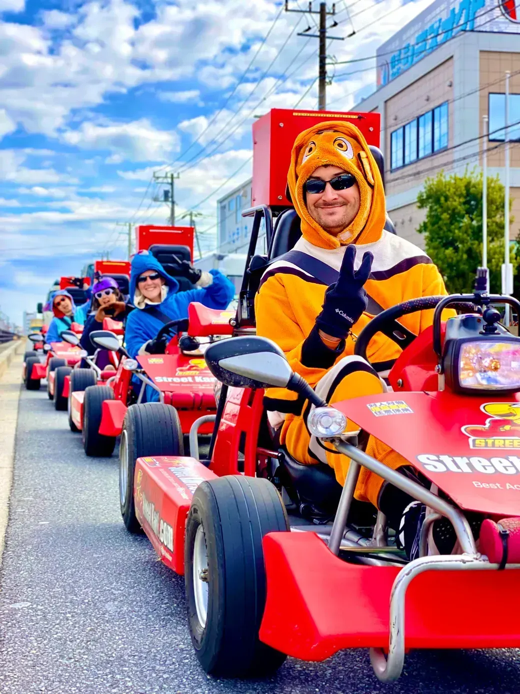 Person in an orange animal hooded costume drives a red go-kart in a street parade, making a peace sign; riders in colorful outfits follow behind.
