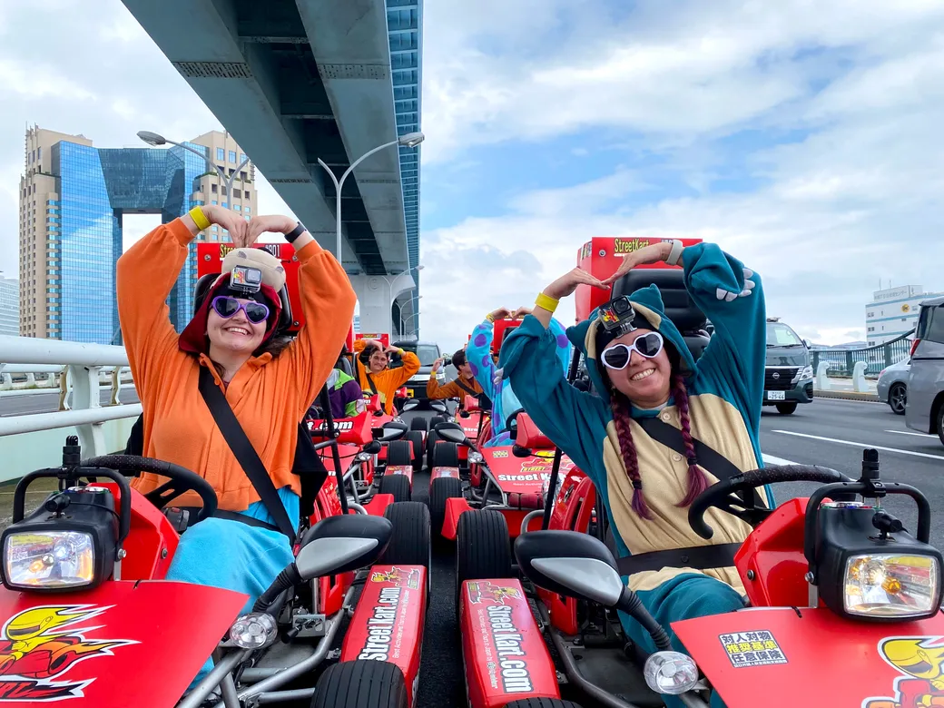 Group of people in orange and teal jumpsuits on red go-karts on a bridge, forming heart shapes with their arms.